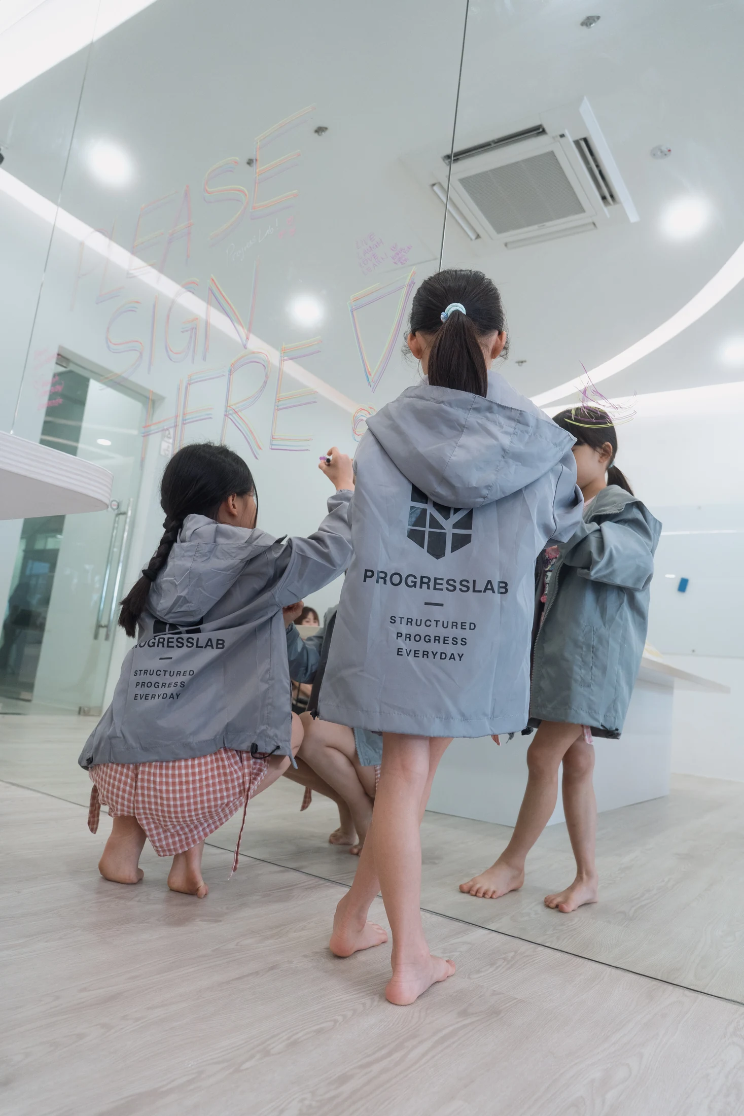 Children in ProgressLab windbreakers drawing together on the centre’s glass wall